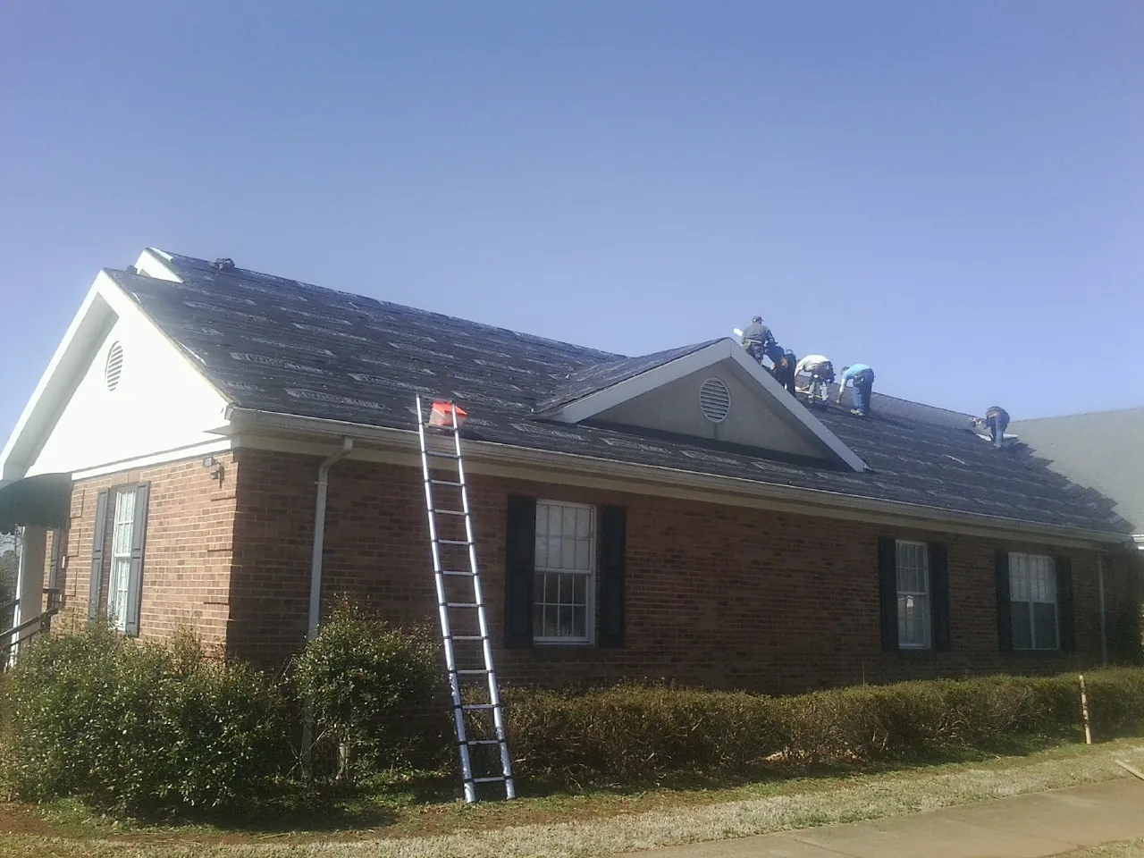 Crew tearing shingles off a brick ranch roof with a ladder set — Metal Roof Repair work by our crew in Brierfield
