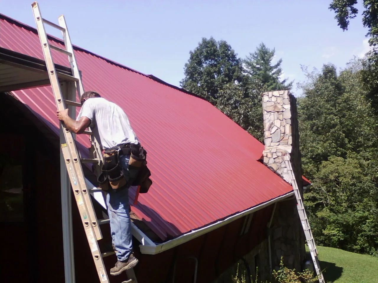 Roofer on a ladder beside a red metal roof and stone chimney — Soffit & Fascia Repair by Roofing Brothers in Brierfield